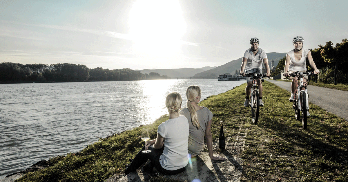 Im Sonnenlicht glänzende Donau: Zwei Radfahrer:innen erkunden Donauradweg in Niederösterreich. (© Donau Niederösterreich/Steve Haider)